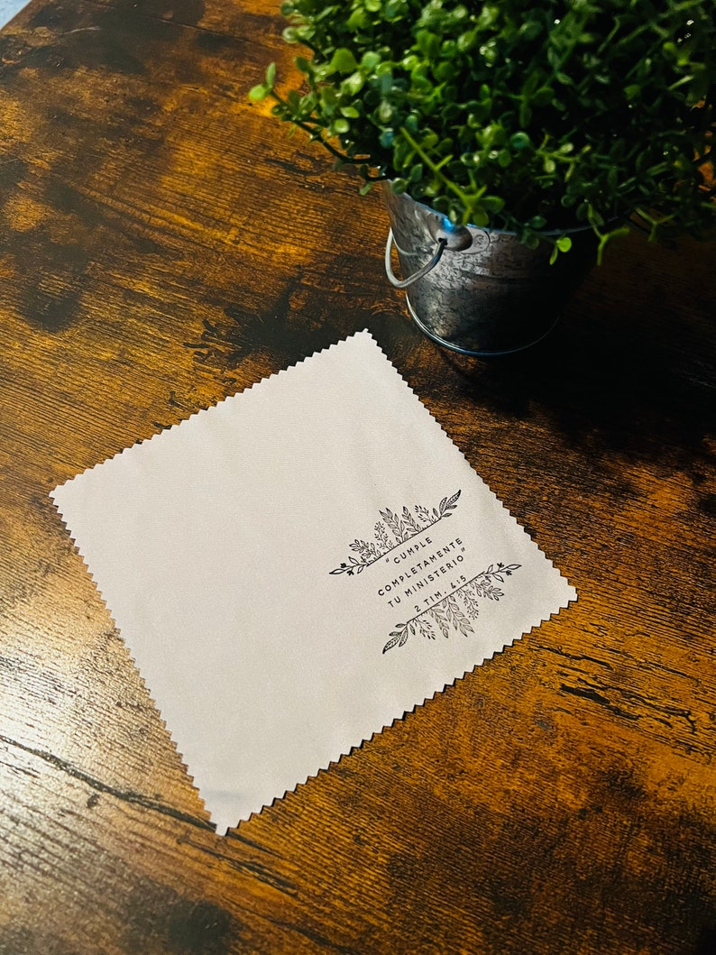 May include: A white microfiber cleaning cloth with a black floral design and the text "Cumple Completamente Tu Ministerio" 2 Tim. 4:5. The cloth is on a brown wooden surface.