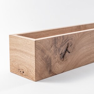 May include: A rectangular wooden planter box with a natural wood finish. The planter has a knot in the wood on the front side.
