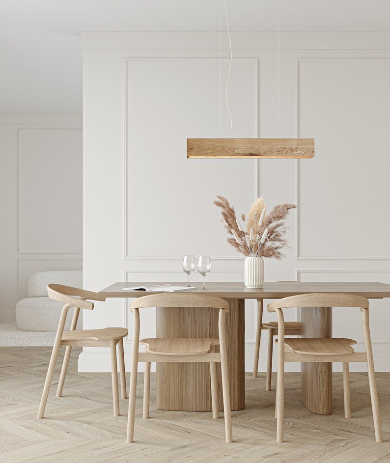 May include: A wooden dining table with four chairs and a wooden rectangular pendant light hanging above. The table and chairs are made of light wood and the pendant light is a warm brown color. There is a vase of dried flowers on the table.