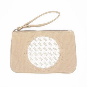 May include: A beige canvas wristlet with a zipper closure and a small handle. A circular patch on the front features the text "3M" repeated in a pattern. The wristlet is a neutral color and appears to be made of a durable material.