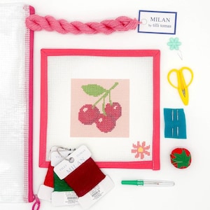 May include: A pink needlepoint kit featuring a cherry design. The kit includes a canvas, embroidery floss, needles, scissors, and a needle threader. The kit is labeled "MILAN by tilli tomas".