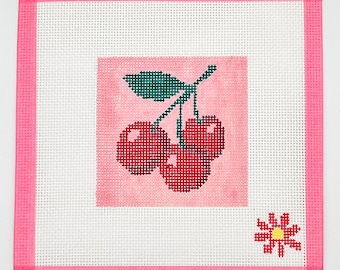 May include: A cross-stitch design featuring three red cherries with green leaves on a pink background. The design is framed with a pink border and includes a small flower in the lower right corner. The artwork is on a white canvas.