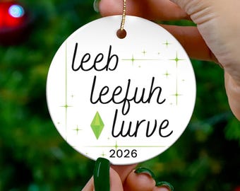 May include: A round white ceramic Christmas ornament featuring the black text leeb leefuh lurve, a green diamond-shaped icon, and the year 2026. The ornament is decorated with small green star accents and hangs from a gold-coloured string.