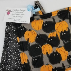 May include: Two black and orange Halloween-themed pot holders with a black cat and pumpkin pattern. The pot holders are reversible with one side featuring a black background with white stars and the other side featuring a gray background with black cats and orange pumpkins.