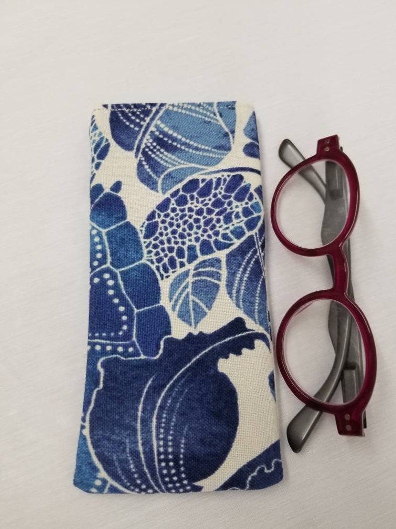 K&ouml;nnte beinhalten: Ein Brillenetui aus blau-wei&szlig;em Stoff mit einer roten und grauen Lesebrille daneben.