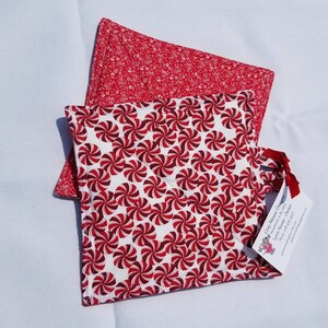May include: Two red and white quilted hot pads. The top pad has a small floral pattern. The bottom pad has a peppermint swirl pattern. The bottom pad has a tag attached that says "Cozy Mouse Designs Handmade in the USA"