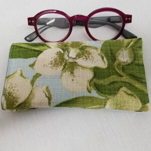 K&ouml;nnte beinhalten: Eine Brillenh&uuml;lle mit floralem Muster auf hellblauem Hintergrund und gr&uuml;nen Bl&auml;ttern. Die H&uuml;lle ist geschlossen und ein Paar burgunderfarbene Lesebrillen liegen darauf.