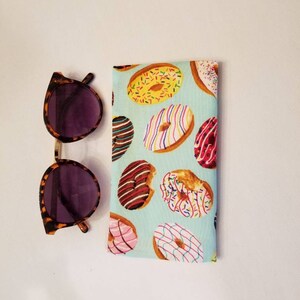 K&ouml;nnte beinhalten: Ein hellblaues Stoff-Sonnenbrillenetui mit einem Donut-Muster. Das Etui ist gef&uuml;llt mit bunten Donuts mit Streuseln, Schokolade und rosa Zuckerguss.