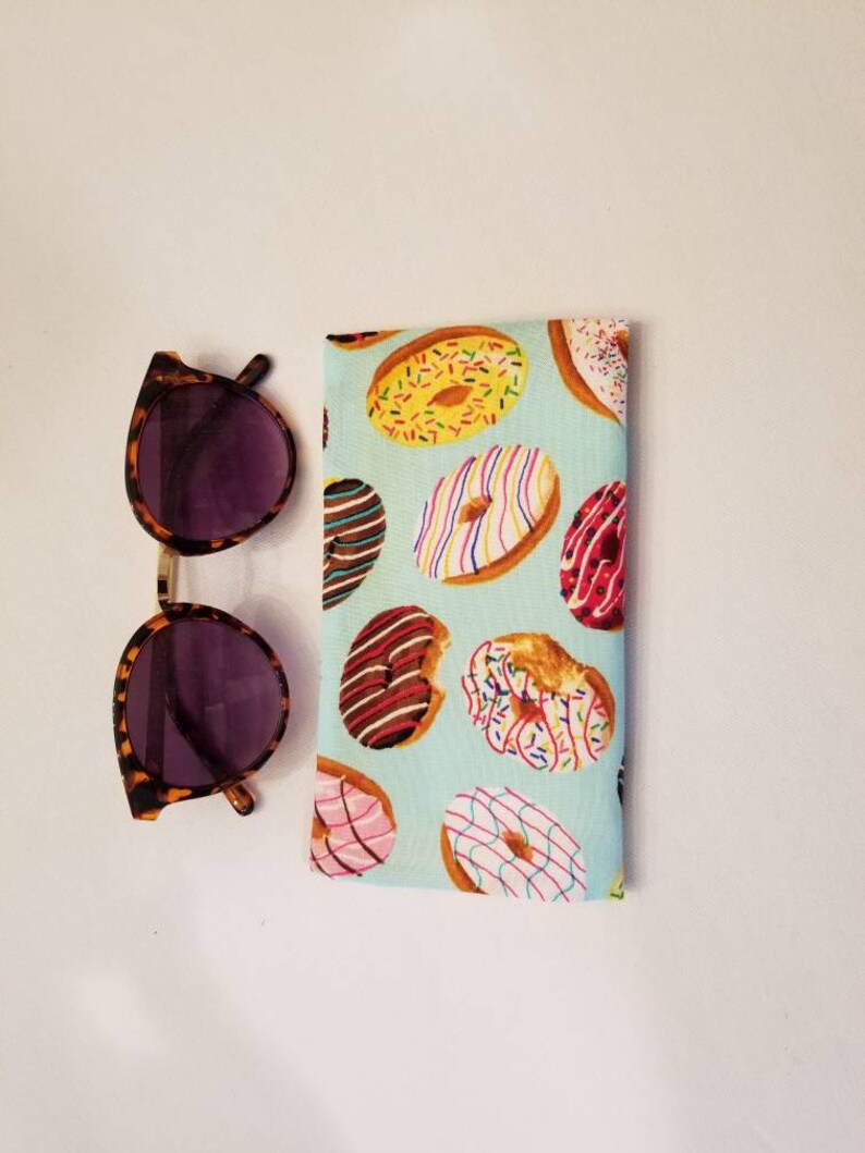 K&ouml;nnte beinhalten: Ein hellblaues Stoff-Sonnenbrillenetui mit einem Donut-Muster. Das Etui ist gef&uuml;llt mit bunten Donuts mit Streuseln, Schokolade und rosa Zuckerguss.