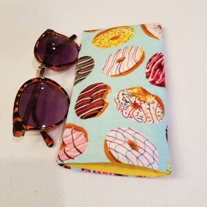 K&ouml;nnte beinhalten: Ein hellblaues Sonnenbrillenetui aus Stoff mit gelber Innenausstattung. Das Etui ist mit einem Muster aus bunten Donuts mit Streuseln, Zuckerguss und Schokolade bedeckt.