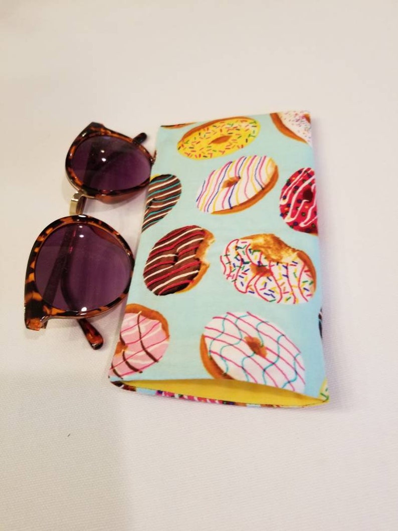 K&ouml;nnte beinhalten: Ein hellblaues Sonnenbrillenetui aus Stoff mit gelber Innenausstattung. Das Etui ist mit einem Muster aus bunten Donuts mit Streuseln, Zuckerguss und Schokolade bedeckt.