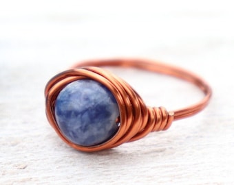 May include: A copper wire-wrapped ring featuring a round, blue-grey gemstone. The ring's band and the wire wrapping are a warm copper color, creating a contrast with the stone. The ring is set against a light background.