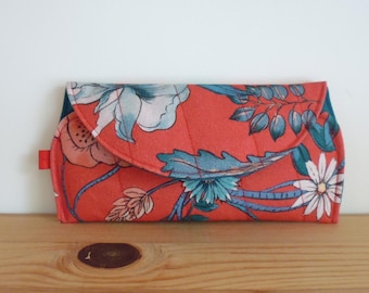 May include: A rectangular fabric clutch with a rounded flap closure, featuring a vibrant coral-orange background printed with a botanical pattern of large white and teal flowers, green leaves, and thin stems, finished with a small red fabric side tab.