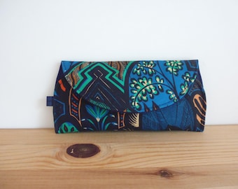 May include: A rectangular clutch with a flap closure, featuring a vibrant African print. The fabric showcases a mix of blue, green, orange, and black geometric and floral patterns. A small blue tab is on the side.