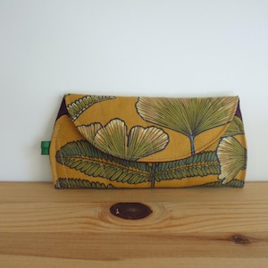 May include: A rectangular fabric clutch with a mustard yellow background featuring a repeating pattern of green ginkgo leaves and fern fronds. The clutch has a flap closure with a curved edge and a small green fabric tab on the left side.