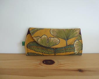 May include: A rectangular fabric clutch with a mustard yellow background featuring a repeating pattern of green ginkgo leaves and fern fronds. The clutch has a flap closure with a curved edge and a small green fabric tab on the left side.