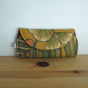 Peut inclure: Une pochette enveloppe en tissu rectangulaire avec un fond jaune moutarde orné d'un motif répétitif de feuilles de ginkgo vertes et dorées avec des accents brun foncé. Le rabat est arrondi et présente une petite languette violet clair.