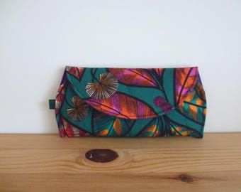 May include: A rectangular fabric clutch bag featuring a vibrant teal background with a repeating pattern of stylised leaves in shades of orange, magenta, and dark brown. The bag has a fold-over flap closure and a small dark green side tab.