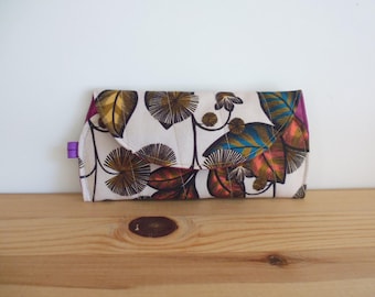 May include: A rectangular fabric clutch with a fold-over flap, featuring a cream-colored background printed with a botanical pattern of stylized leaves in shades of brown, teal, and orange, accented by a small purple tab on the left side.