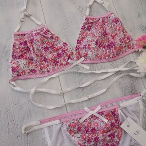 May include: A floral print lingerie set featuring a triangle bra and matching thong. The set is adorned with a pink floral pattern on a white background, white straps, and delicate lace trim. A small tag is attached.