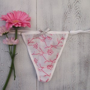 May include: White thong underwear with a floral pattern in pink, accented with a small gray bow. The lingerie is displayed with pink gerbera daisies and other pink flowers against a white wooden background.