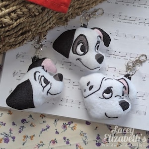 Puede incluir: Tres llaveros de felpa con forma de cabeza de perro dálmata, con marcas en blanco y negro y orejas rosas. Cada llavero tiene un cierre plateado. Los llaveros están sobre una partitura con el texto "Cruella De".
