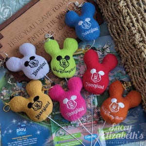 May include: A selection of Mickey Mouse-shaped keyrings in various colours. Each plush keyring features the iconic mouse face and the Disneyland or Walt Disney World logo. The keyrings are in shades of blue, red, yellow, pink, white, green, and orange.