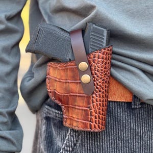 May include: A brown leather holster with a crocodile pattern and two brass snaps. A black handgun is partially visible inside the holster.