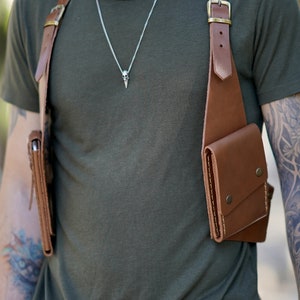 May include: A brown leather chest rig with two pockets, worn over a green t-shirt. The rig has a buckle on the shoulder strap and a metal snap closure on the pockets.