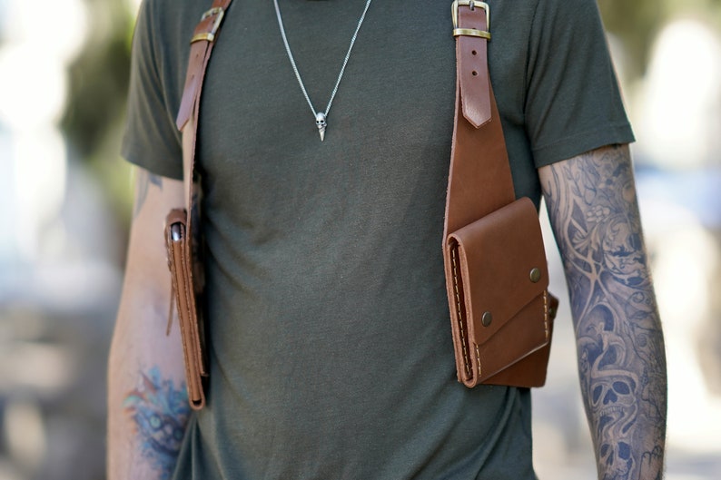 May include: A brown leather chest rig with two pockets, worn over a green t-shirt. The rig has a buckle on the shoulder strap and a metal snap closure on the pockets.
