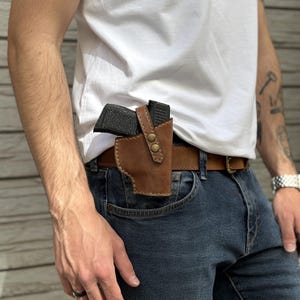 Könnte beinhalten: Ein braunes Lederholster mit einer Waffe darin, befestigt an einem braunen Gürtel. Das Holster ist mit zwei messingfarbenen Druckknöpfen befestigt. Der Gürtel wird mit einer blauen Jeans und einem weißen T-Shirt getragen.