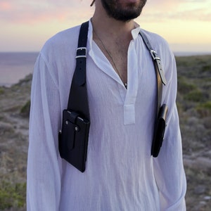May include: A black leather chest rig with two pouches, worn over a white shirt. The rig has adjustable straps and silver buckles.