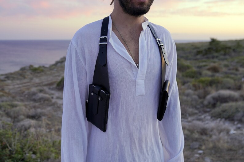 May include: A black leather chest rig with two pouches, worn over a white shirt. The rig has adjustable straps and silver buckles.