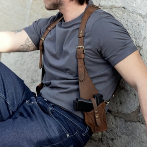 May include: Brown leather shoulder holster with a gun. The holster has adjustable straps and a buckle closure. The gun is black. The holster is worn over a gray t-shirt and blue jeans.