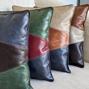 Könnte beinhalten: Vier dekorative Kissen mit Patchwork-Muster. Jedes Kissen besteht aus verschiedenen Lederstücken in verschiedenen Farben, darunter Braun, Grün, Rot und Blau. Die Kissen sind in einer Reihe auf einem hellbeigen Sofa angeordnet.