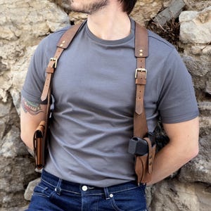 May include: A brown leather shoulder holster with two pouches and adjustable straps. The holster has gold-colored buckles and is designed to be worn across the chest and back. The leather appears to be distressed.