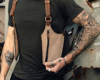 May include: A brown leather chest rig with a zippered pocket and adjustable straps. The rig is worn by a person with tattoos on their arms.