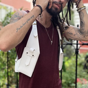 May include: White leather crossbody bag with a strap that goes over the shoulder. The bag has a flap with a buckle closure and a small pocket on the front. The bag is worn by a person with tattoos on their arms.