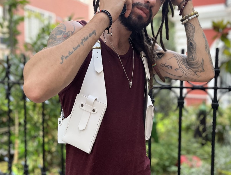 May include: White leather crossbody bag with a strap that goes over the shoulder. The bag has a flap with a buckle closure and a small pocket on the front. The bag is worn by a person with tattoos on their arms.