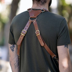 May include: A brown leather camera strap with brass buckles and a strap that goes over the chest. The strap is worn by a person with a tattoo on their arm.