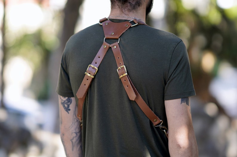 May include: A brown leather camera strap with brass buckles and a strap that goes over the chest. The strap is worn by a person with a tattoo on their arm.