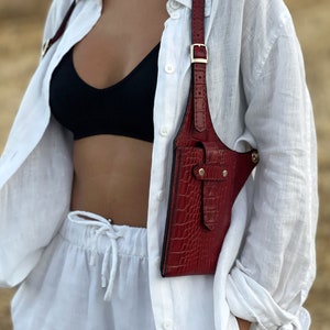 May include: A red leather crossbody bag with a crocodile-embossed design. The bag has a strap that goes over the shoulder and a buckle closure. The bag is worn over a white shirt and white pants.