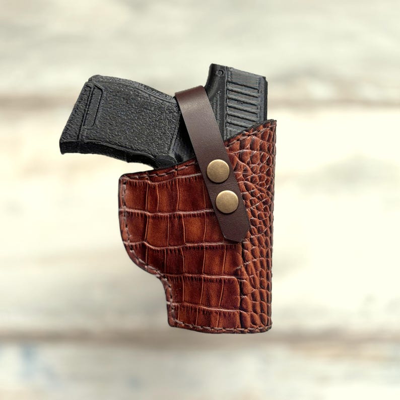 May include: A brown leather holster with a crocodile pattern and two brass snaps. A black handgun is inside the holster.