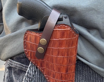 May include: A brown leather holster with a crocodile pattern and a black handgun inside. The holster has a brown leather strap with two brass snaps.