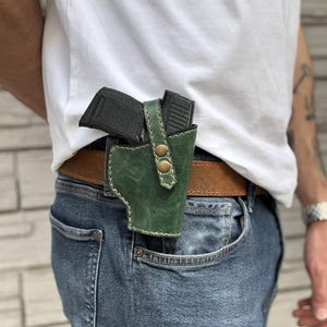 May include: A green leather holster with a black handgun inside, attached to a brown belt. The holster has brass-colored rivets and is worn on the hip with blue jeans.