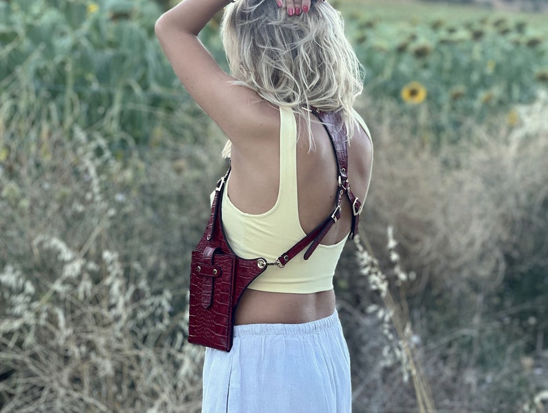 May include: A red leather crossbody bag with a strap that goes over the shoulders. The bag has a flap closure and a small pocket on the front. The bag is worn by a person in a yellow tank top and white pants.