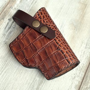 May include: A brown leather holster with a crocodile pattern. The holster has a flap with two brass snaps.