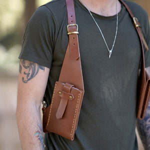 May include: A brown leather crossbody bag with two pockets. The bag has a strap that goes over the shoulder and a buckle on the strap. The bag is worn by a person with tattoos on their arms.