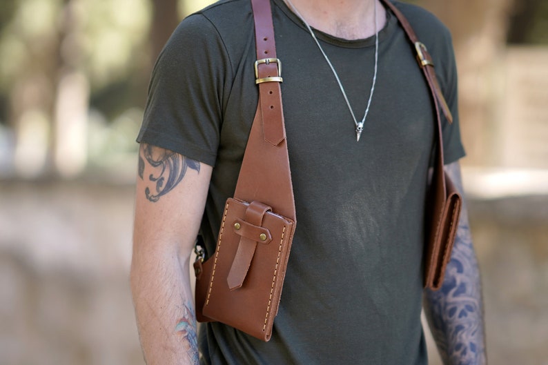 May include: A brown leather crossbody bag with two pockets. The bag has a strap that goes over the shoulder and a buckle on the strap. The bag is worn by a person with tattoos on their arms.