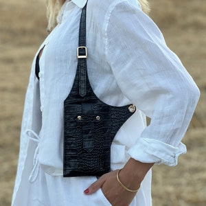 May include: A black leather crossbody bag with a crocodile-embossed design. The bag has a gold buckle and a gold chain strap.